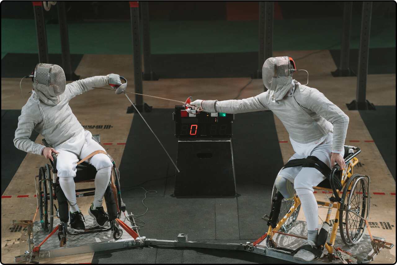 Two PWD athletes engaged in a fencing match, wearing full fencing gear and protective masks.